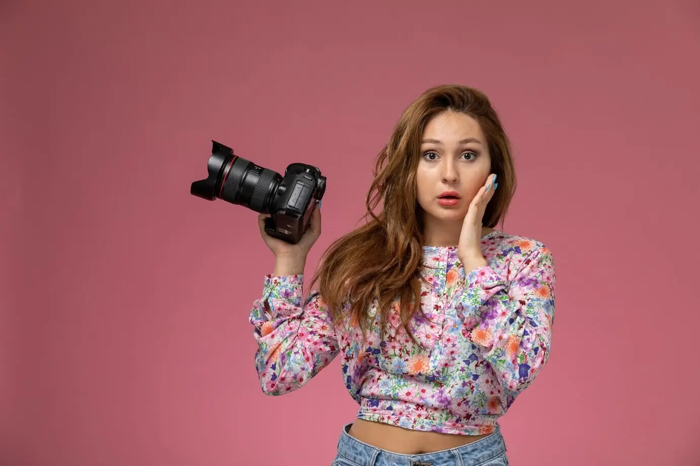 Vorderansicht einer jungen, schönen Frau in blumengemustertem Shirt und blauen Jeans, die eine Fotokamera hält, vor pinkem Hintergrund.